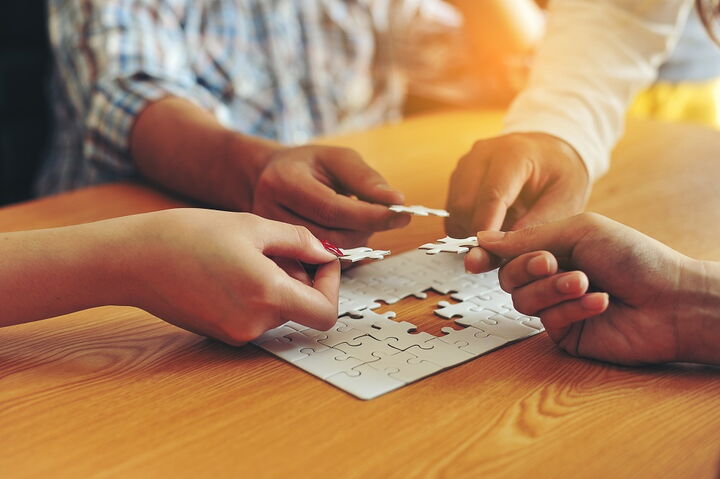 vier Hände die jeweils ein Puzzleteil halten und zu einem Gesamtpuzzle einfügen