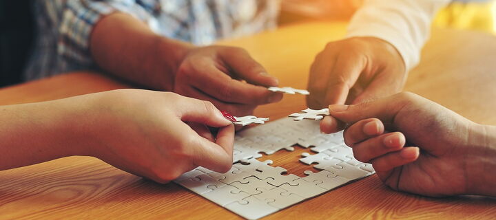 vier Hände die jeweils ein Puzzleteil halten und zu einem Gesamtpuzzle einfügen